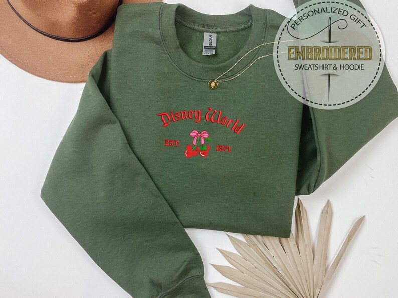 May include: An olive green sweatshirt with the embroidered text "Disney World" in red script, with a pink bow and cherries design. The sweatshirt also has the text "EST. 1971". A gold necklace and a brown hat are also visible.