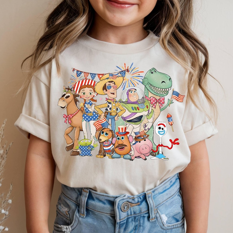 Puede incluir: Una camiseta de color crema con un estampado gr&aacute;fico de personajes de Toy Story, incluidos Woody, Buzz Lightyear, Jessie, Rex y Forky. Los personajes est&aacute;n decorados con motivos de banderas estadounidenses, estrellas, rayas y fuegos artificiales.