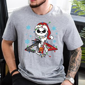 Puede incluir: Una camiseta gris con un gr&aacute;fico navide&ntilde;o de Jack Skellington de Pesadilla antes de Navidad. Lleva un gorro de Pap&aacute; Noel y est&aacute; rodeado de luces navide&ntilde;as.