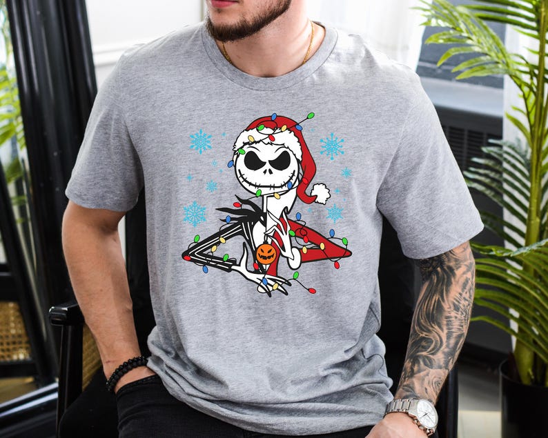 Puede incluir: Una camiseta gris con un gr&aacute;fico navide&ntilde;o de Jack Skellington de Pesadilla antes de Navidad. Lleva un gorro de Pap&aacute; Noel y est&aacute; rodeado de luces navide&ntilde;as.