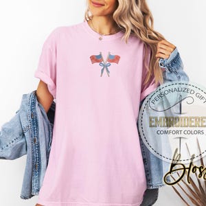 May include: A light pink t-shirt with an embroidered design of two crossed American flags tied with a blue bow. The shirt has rolled-up sleeves and is part of the Comfort Colors collection. The image also includes the text "PERSONALIZED GIFT" and "EMBROIDERED".