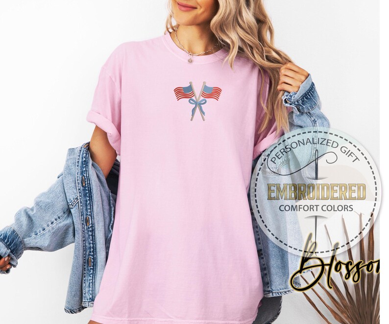 May include: A light pink t-shirt with an embroidered design of two crossed American flags tied with a blue bow. The shirt has rolled-up sleeves and is part of the Comfort Colors collection. The image also includes the text "PERSONALIZED GIFT" and "EMBROIDERED".
