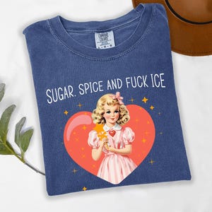 May include: A navy blue t-shirt with the text "SUGAR. SPICE AND FUCK ICE". The graphic features a vintage illustration of a girl holding a star wand inside a heart. The shirt is a Comfort Colors brand.