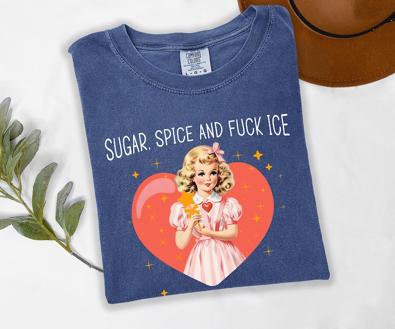May include: A navy blue t-shirt with the text "SUGAR. SPICE AND FUCK ICE". The graphic features a vintage illustration of a girl holding a star wand inside a heart. The shirt is a Comfort Colors brand.