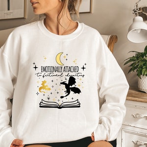 May include: White sweatshirt with a black and gold design. The design features a crescent moon, stars, two dragons, and an open book. The text on the sweatshirt reads "Emotionally attached to fictional characters."