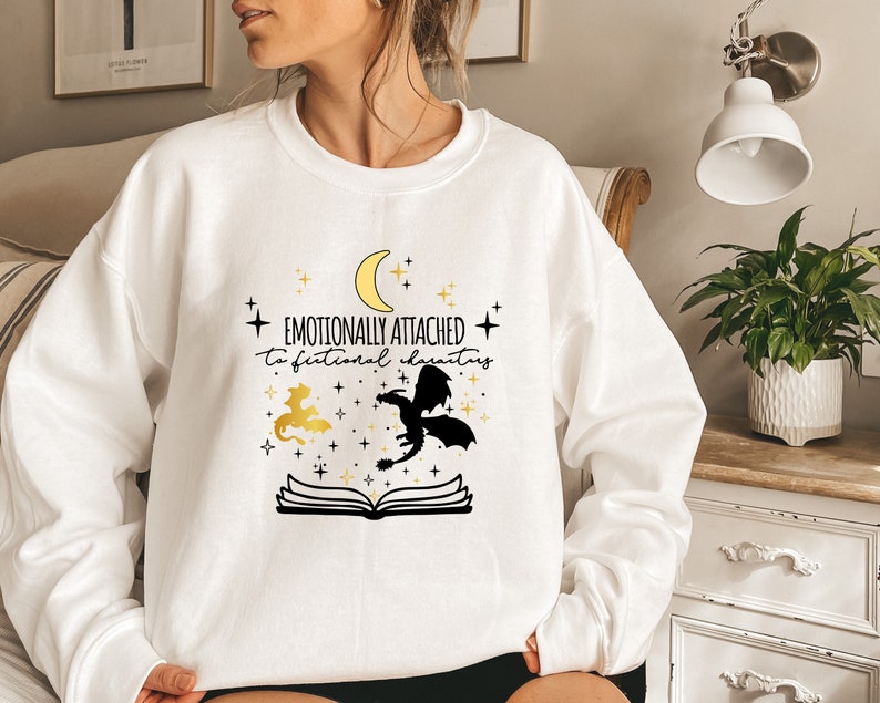 May include: White sweatshirt with a black and gold design. The design features a crescent moon, stars, two dragons, and an open book. The text on the sweatshirt reads "Emotionally attached to fictional characters."