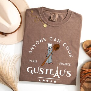 May include: A brown t-shirt with the slogan "Anyone Can Cook" and a cartoon rat holding a wooden spoon. The shirt also features the text "Paris France" and "GUSTEAU'S". A gold necklace, sandals, and a hat are also in the image.