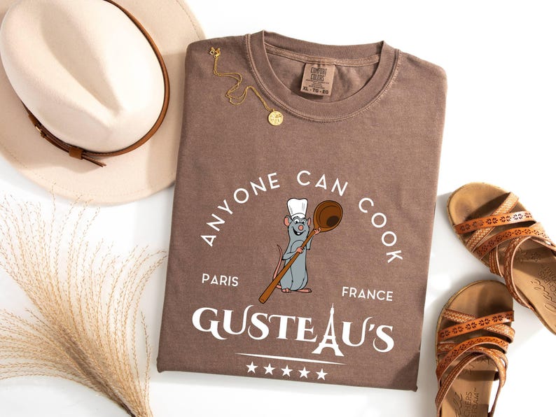 May include: A brown t-shirt with the slogan "Anyone Can Cook" and a cartoon rat holding a wooden spoon. The shirt also features the text "Paris France" and "GUSTEAU'S". A gold necklace, sandals, and a hat are also in the image.