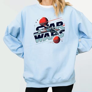Puede incluir: Sudadera azul claro con un gr&aacute;fico de una nave espacial volando por el espacio con el texto "STAR WARS" en un estilo retro.