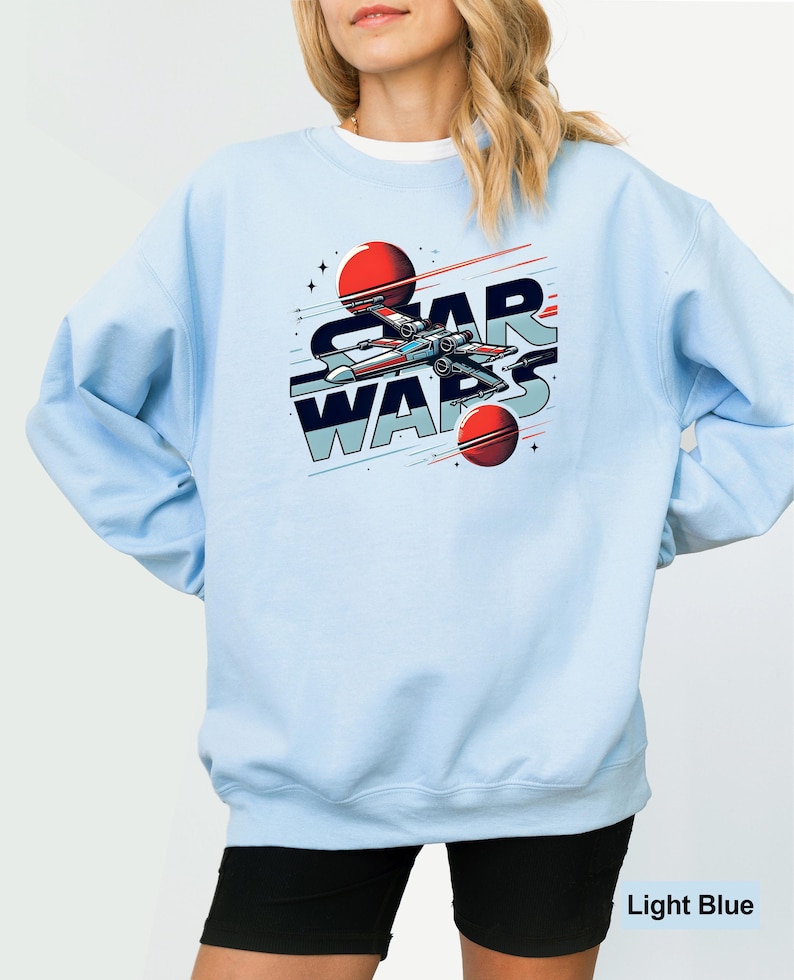 Puede incluir: Sudadera azul claro con un gr&aacute;fico de una nave espacial volando por el espacio con el texto "STAR WARS" en un estilo retro.