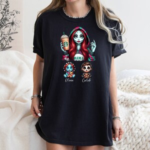 May include: Black t-shirt with a graphic print of a woman with red hair and a green and black dress holding a coffee cup. The woman is surrounded by three smaller figures, two of which are labeled "Olivia" and "Caleb". The text "MAMA" is above the figures.