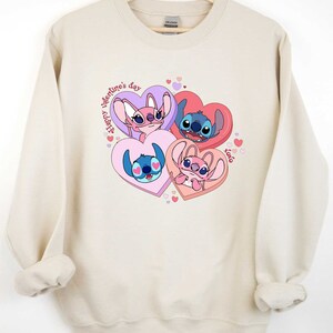 Puede incluir: Una sudadera beige con un estampado gr&aacute;fico de Stitch y Angel de Lilo & Stitch. El gr&aacute;fico presenta a los personajes en corazones con el texto "Happy Valentine's Day" y "xoxo".