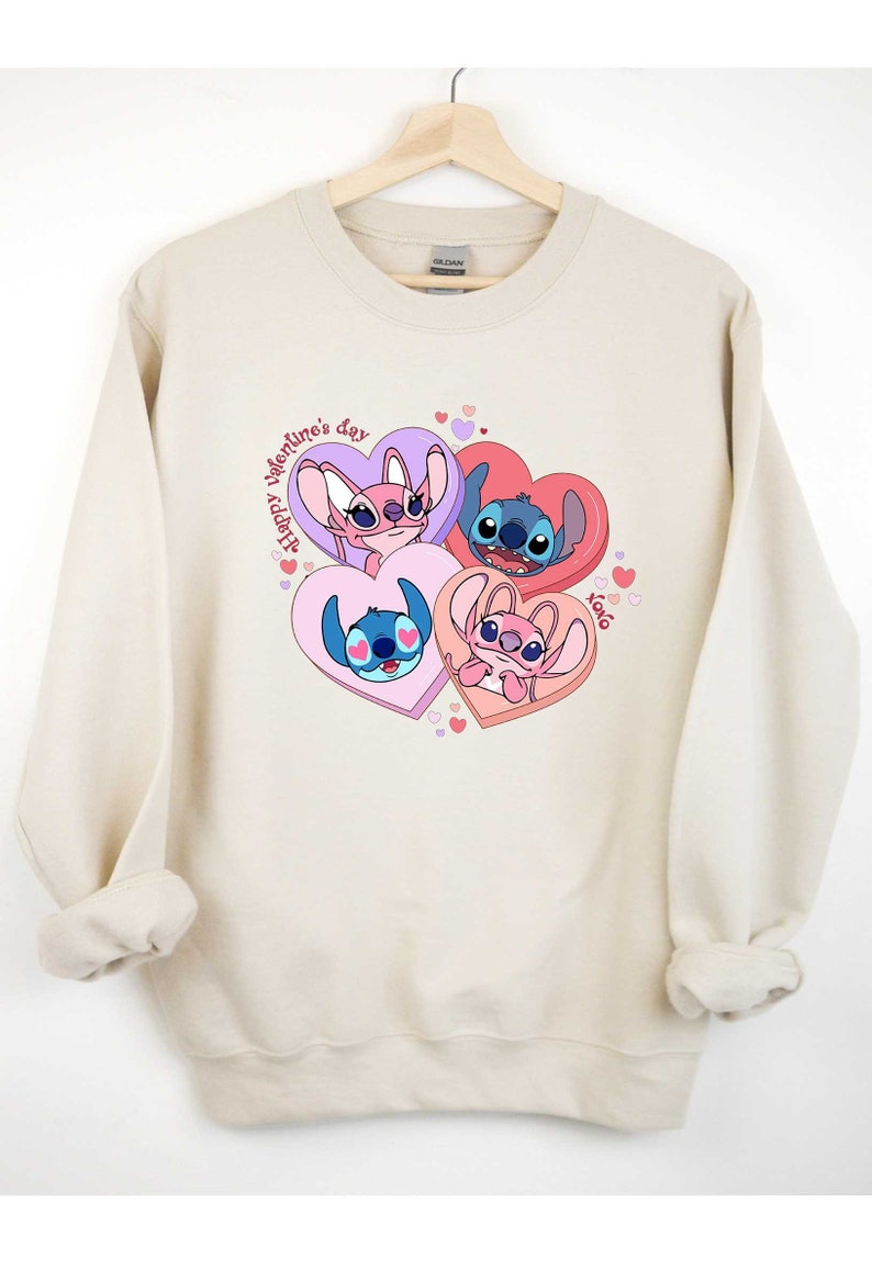 Puede incluir: Una sudadera beige con un estampado gr&aacute;fico de Stitch y Angel de Lilo & Stitch. El gr&aacute;fico presenta a los personajes en corazones con el texto "Happy Valentine's Day" y "xoxo".