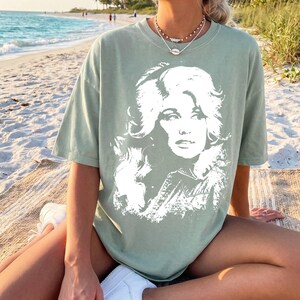 May include: A sage green short-sleeve t-shirt featuring a large, white, high-contrast graphic print of a woman with voluminous hair on the front. The shirt has a crew neckline and a relaxed, loose-fitting silhouette with a standard hemline.
