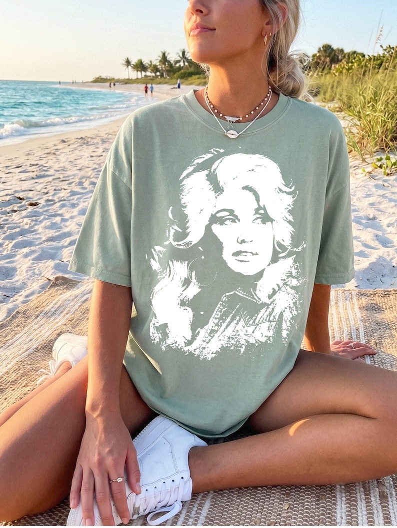 May include: A sage green short-sleeve t-shirt featuring a large, white, high-contrast graphic print of a woman with voluminous hair on the front. The shirt has a crew neckline and a relaxed, loose-fitting silhouette with a standard hemline.