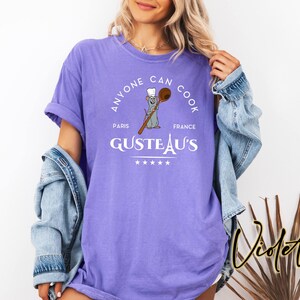 May include: A violet t-shirt featuring the text "ANYONE CAN COOK" in a circular design. The design includes a cartoon rat holding a wooden spoon, the words "PARIS FRANCE" and "GUSTEAU'S".
