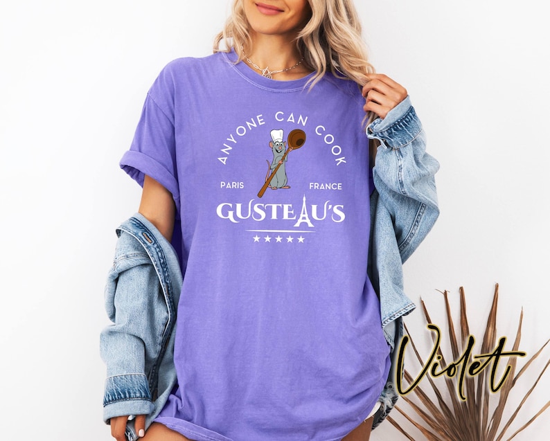May include: A violet t-shirt featuring the text "ANYONE CAN COOK" in a circular design. The design includes a cartoon rat holding a wooden spoon, the words "PARIS FRANCE" and "GUSTEAU'S".
