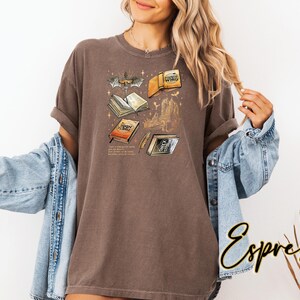 May include: Espresso-coloured t-shirt with a graphic of open books, a bat, and a castle. The text on the shirt includes the words "Iron Flame" and other text. The shirt is worn with a blue denim jacket.