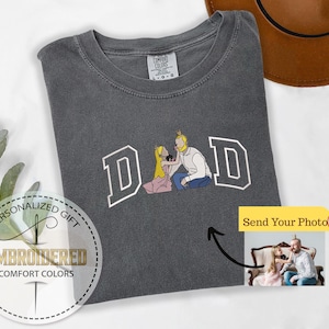 May include: Dark gray Comfort Colors t-shirt with an embroidered design. The design features the word "DAD" in white block letters with an illustration of a father and two children. The shirt also has a "Send Your Photo" button.