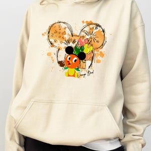 May include: Cream-colored hoodie featuring a graphic of the Orange Bird character with a Mickey Mouse ear design. The design includes orange and yellow accents, with the text "Orange Bird" below the character. The hoodie has a front pocket.