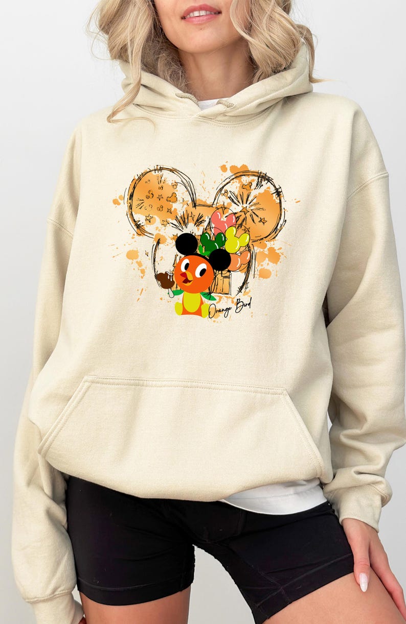 May include: Cream-colored hoodie featuring a graphic of the Orange Bird character with a Mickey Mouse ear design. The design includes orange and yellow accents, with the text "Orange Bird" below the character. The hoodie has a front pocket.