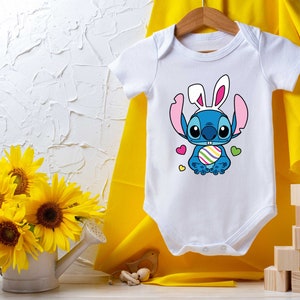 May include: White baby onesie with a blue cartoon character wearing bunny ears and holding an Easter egg. The character has large eyes and a big smile. The onesie is on a yellow background.