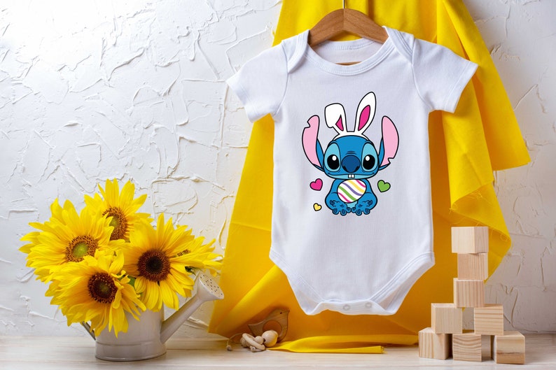 May include: White baby onesie with a blue cartoon character wearing bunny ears and holding an Easter egg. The character has large eyes and a big smile. The onesie is on a yellow background.