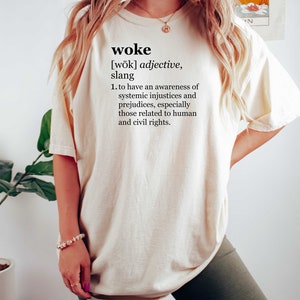 May include: A white t-shirt with the word "woke" and its definition printed on the front. The definition is: "[wōk] adjective, slang 1. to have an awareness of systemic injustices and prejudices, especially those related to human and civil rights."
