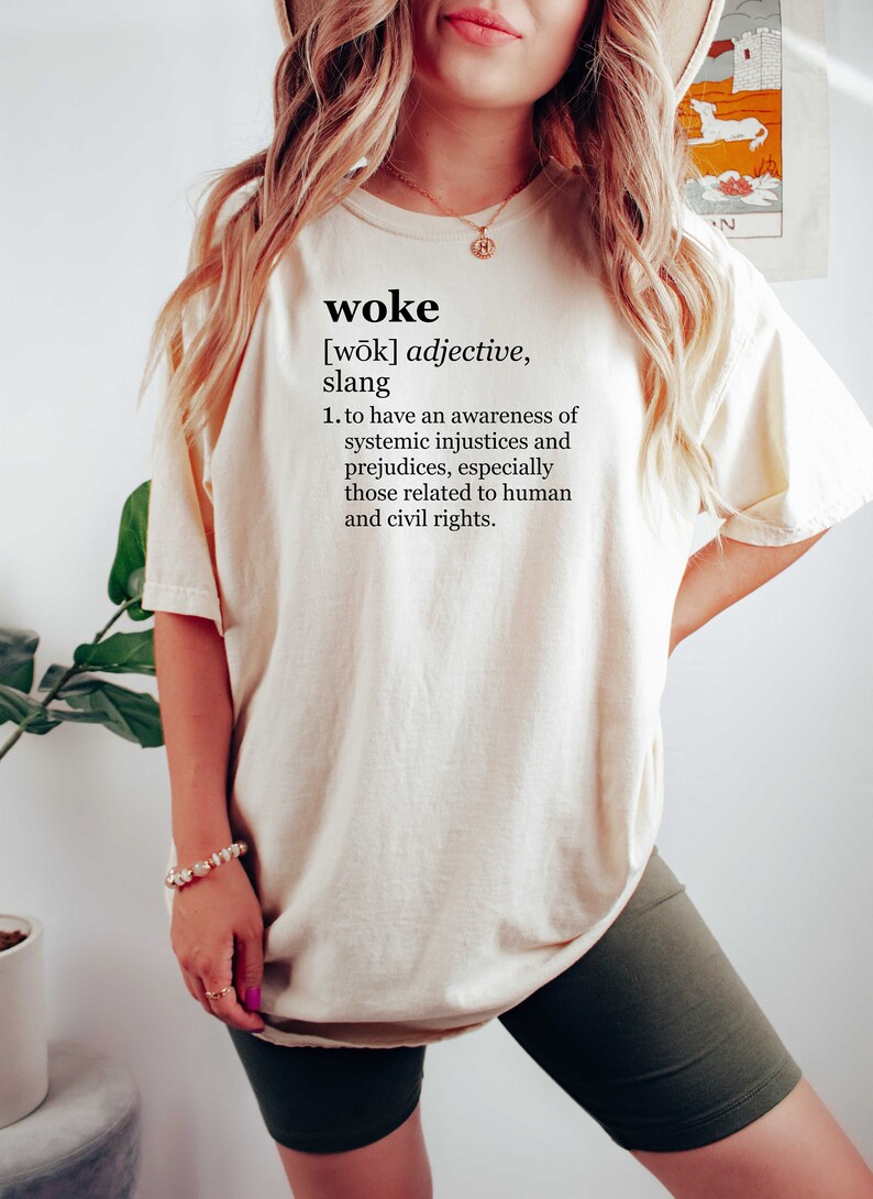 May include: A white t-shirt with the word "woke" and its definition printed on the front. The definition is: "[wōk] adjective, slang 1. to have an awareness of systemic injustices and prejudices, especially those related to human and civil rights."