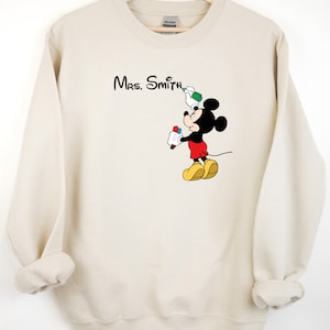 May include: A beige sweatshirt with a Mickey Mouse graphic. Mickey is holding a red marker and a green spray paint can. The text "Mrs. Smith" is printed above Mickey.