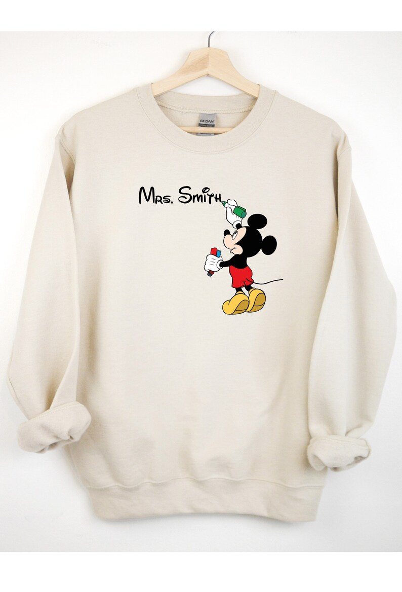May include: A beige sweatshirt with a Mickey Mouse graphic. Mickey is holding a red marker and a green spray paint can. The text "Mrs. Smith" is printed above Mickey.