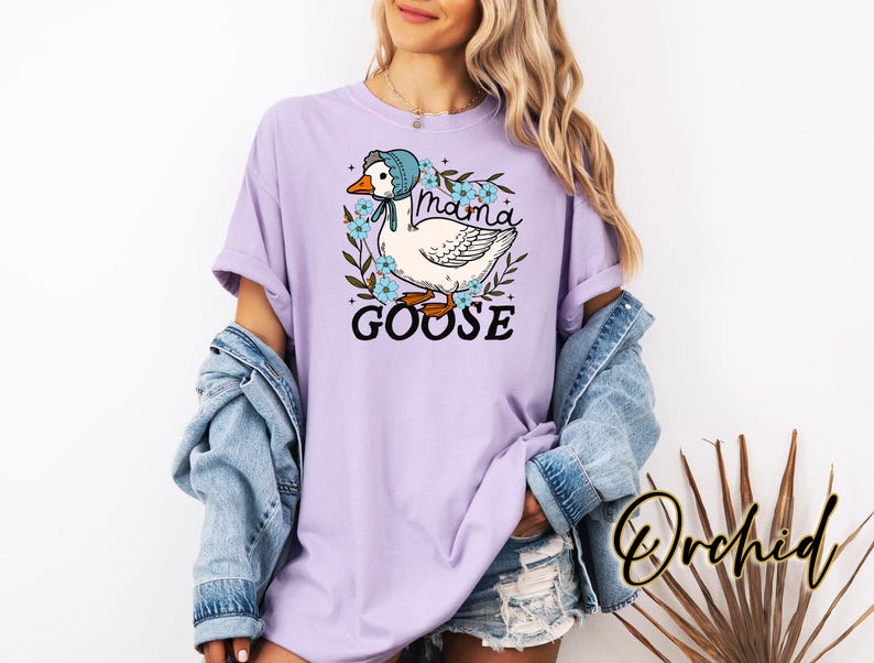 Puede incluir: Una camiseta de color lavanda con un gr&aacute;fico de una oca con un gorro azul y rodeada de flores. El texto "Mama Goose" est&aacute; impreso debajo de la oca.