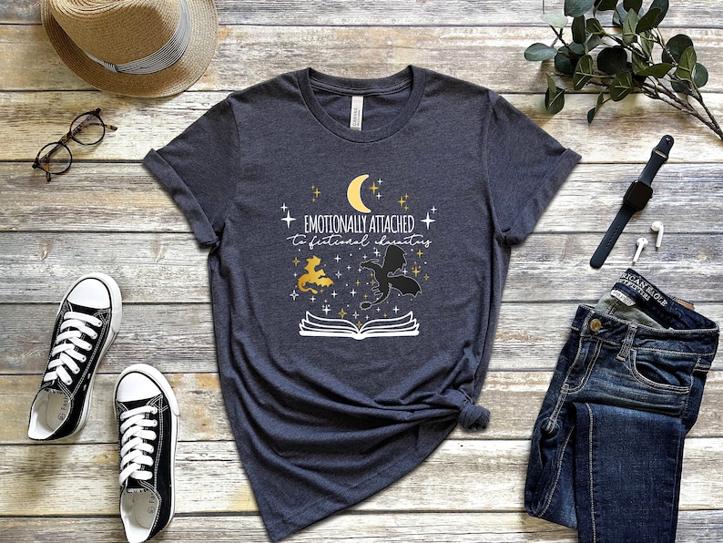 May include: A dark heather grey t-shirt with a graphic of a black dragon and a gold dragon flying over an open book with a crescent moon and stars. The text on the shirt reads "Emotionally Attached to Fictional Characters".