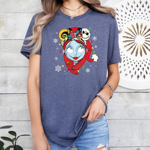 Puede incluir: Una camiseta azul con un gr&aacute;fico de un personaje de Pesadilla Antes de Navidad con un gorro de Pap&aacute; Noel y rodeado de luces de Navidad y copos de nieve.