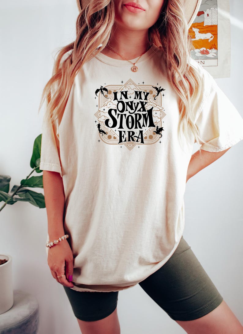 May include: A white t-shirt with a black graphic design that says "In My Onyx Storm Era" with a gold outline and black dragon silhouettes.