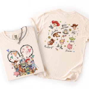 May include: Two cream-coloured t-shirts. One features a Mickey Mouse head design with Disney characters. The other shirt has various Toy Story characters and text. A silver chain necklace is draped over the first shirt.
