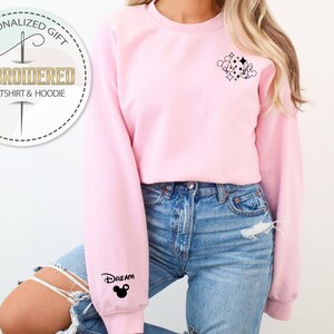 May include: A light pink embroidered sweatshirt with a small black graphic on the upper right chest and the word "Dream" with a Mickey Mouse head on the left sleeve. The sweatshirt has a crew neck and long sleeves. The image also includes a circular logo that says "Embroidered Sweatshirt & Hoodie".