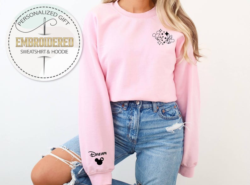 May include: A light pink embroidered sweatshirt with a small black graphic on the upper right chest and the word "Dream" with a Mickey Mouse head on the left sleeve. The sweatshirt has a crew neck and long sleeves. The image also includes a circular logo that says "Embroidered Sweatshirt & Hoodie".