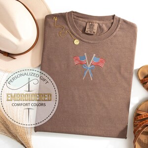 May include: A brown Comfort Colors t-shirt with an embroidered design of two crossed American flags and a blue bow. The shirt is paired with a beige hat, gold necklace, and brown sandals. The shirt has the text "Embroidered Comfort Colors".
