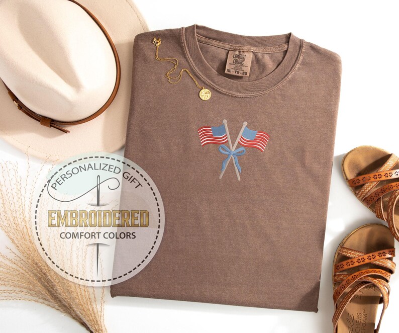 May include: A brown Comfort Colors t-shirt with an embroidered design of two crossed American flags and a blue bow. The shirt is paired with a beige hat, gold necklace, and brown sandals. The shirt has the text "Embroidered Comfort Colors".