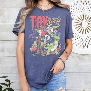 May include: Navy blue t-shirt with a "Toy Story" graphic. The design features Woody, Buzz Lightyear, and Rex the dinosaur, with the text "TOY STORY" in pink and yellow. The text "SINCE 1995" is also included in the design.