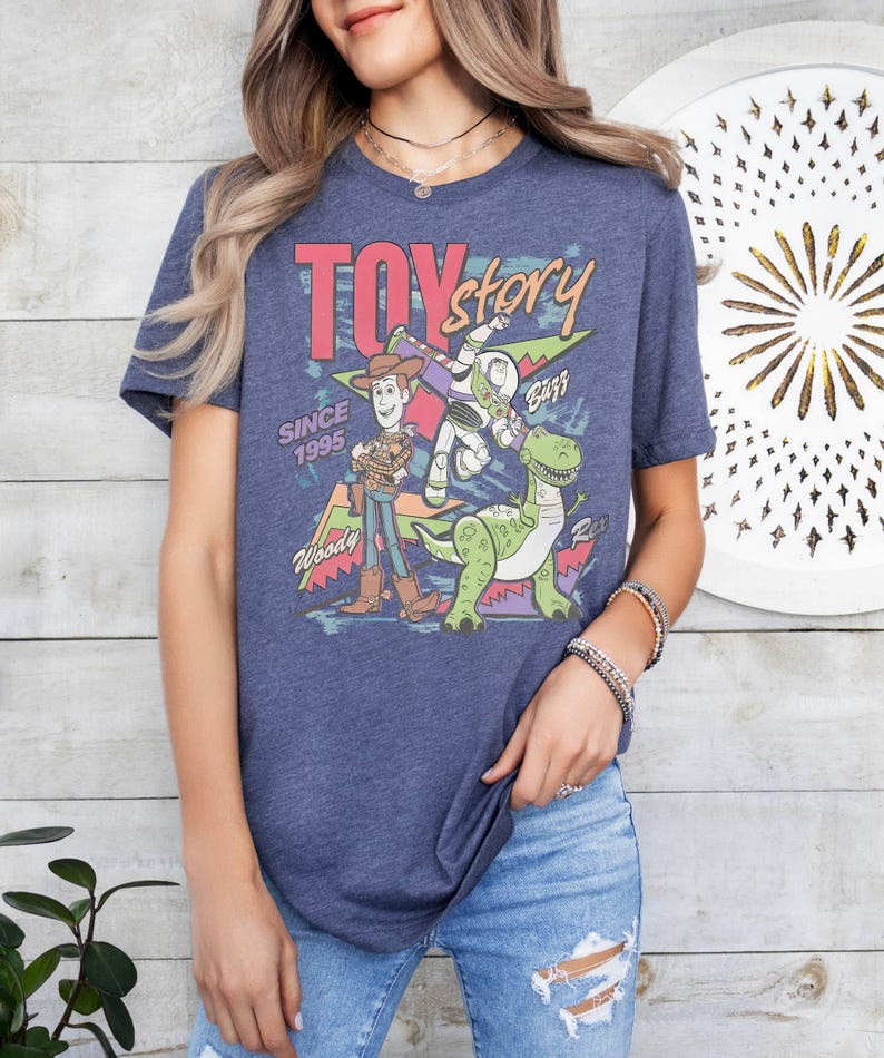 May include: Navy blue t-shirt with a "Toy Story" graphic. The design features Woody, Buzz Lightyear, and Rex the dinosaur, with the text "TOY STORY" in pink and yellow. The text "SINCE 1995" is also included in the design.
