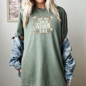 May include: A green t-shirt with a vintage-style graphic that says "In My Onyx Storm Era" with stars and dragons around the text.