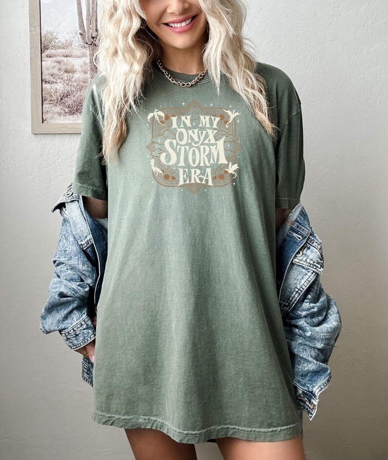 May include: A green t-shirt with a vintage-style graphic that says "In My Onyx Storm Era" with stars and dragons around the text.