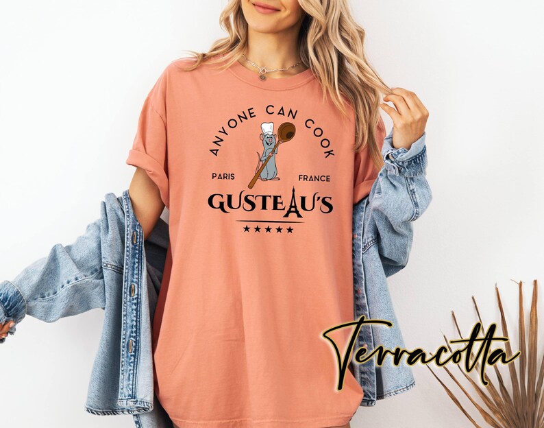 May include: A terracotta-coloured t-shirt featuring the phrase "Anyone Can Cook" in a circular design. The design includes a cartoon rat chef with a wooden spoon, the words "Paris France" and "Gusteau's" with stars.