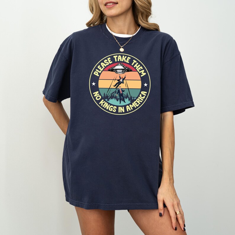 Op de afbeelding: Marineblauwe t-shirt met een cirkelvormige afbeelding. De afbeelding toont een UFO die een persoon ontvoert, met de tekst "PLEASE TAKE THEM, NO KINGS IN AMERICA". Het ontwerp heeft een retro zonsondergang kleurenschema.