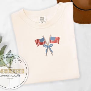 May include: Cream-colored t-shirt with an embroidered design of two crossed American flags tied with a blue bow. The flags have red and white stripes and blue fields with white stars. The shirt is labeled "Comfort Colors" and is a personalized gift.