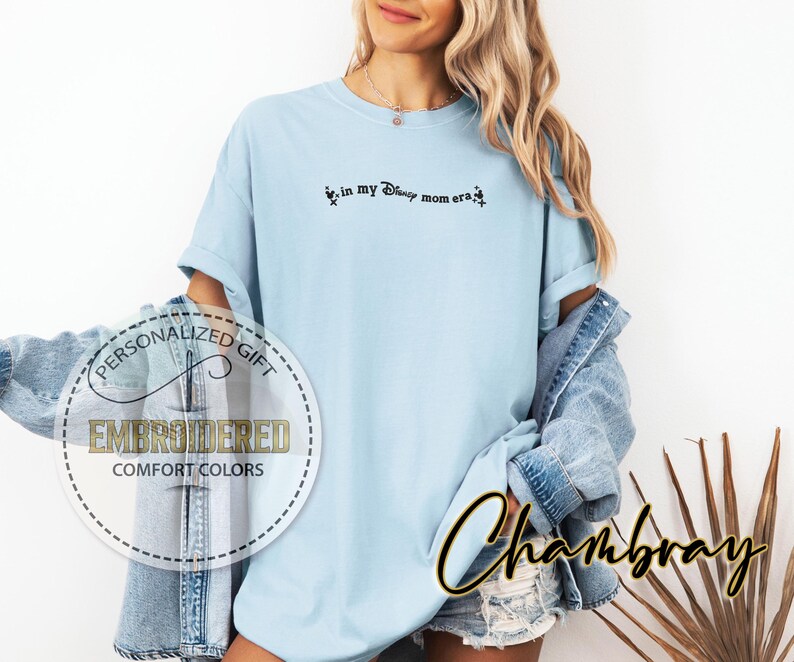 May include: Light blue t-shirt with the text "in my Disney mom era" in black script. The shirt has a relaxed fit and is part of a personalized gift collection. The shirt is made of Comfort Colors material.