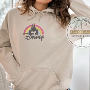 May include: A beige hoodie with a rainbow and castle graphic and the word "DISNEY" embroidered on the front. The hoodie has a drawstring hood and a kangaroo pocket.