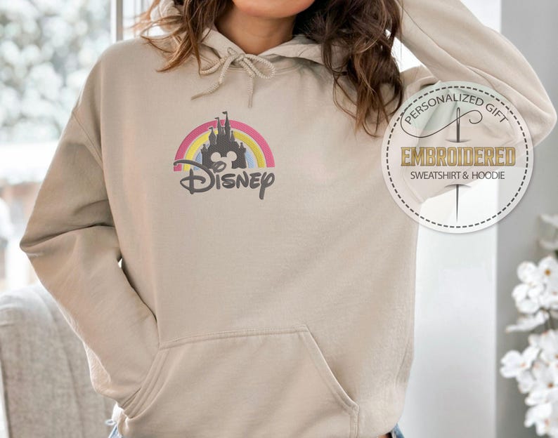 May include: A beige hoodie with a rainbow and castle graphic and the word "DISNEY" embroidered on the front. The hoodie has a drawstring hood and a kangaroo pocket.